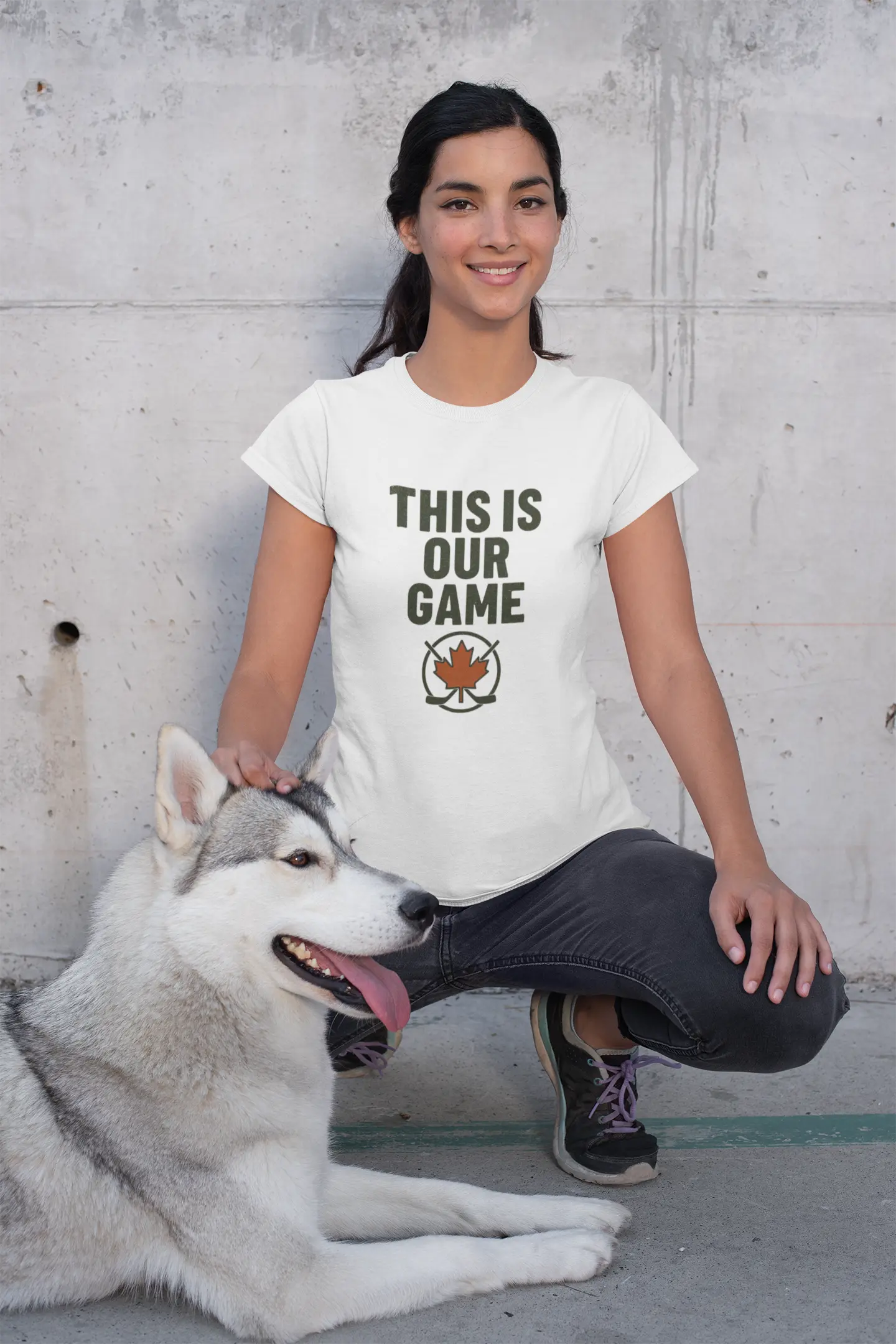 Woman wearing white ‘This Is Our Game’ women’s T-shirt with maple leaf graphic, kneeling next to a husky dog – Maple Stick Hockey product.