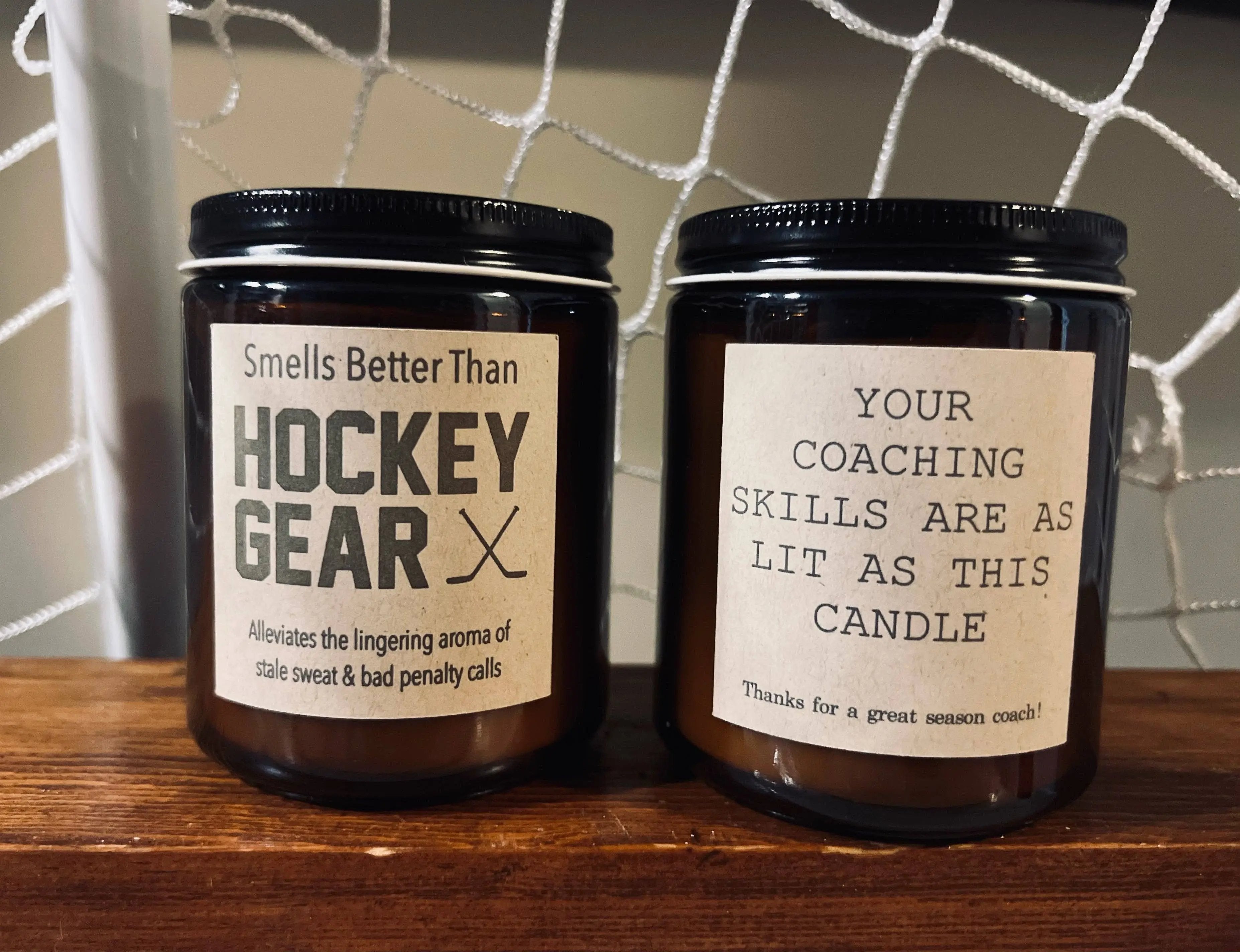 Two black glass candles with humorous labels: one says 'Smells better than hockey gear' and the other says 'Your coaching skills are as lit as this candle' – Maple Stick Hockey product.