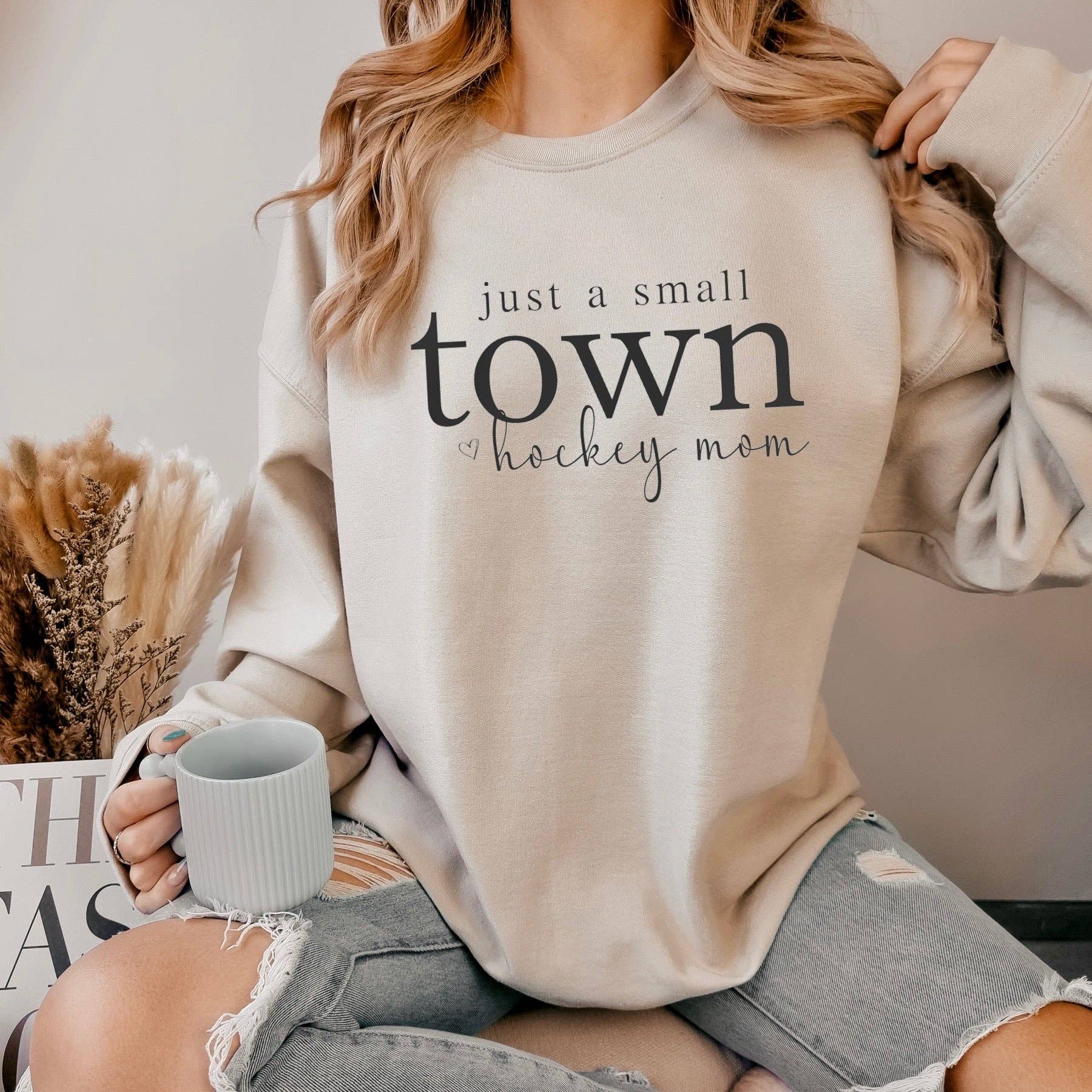 Woman wearing beige "Just a small town hockey mom" sweatshirt holding a grey mug – Maple Stick Hockey product