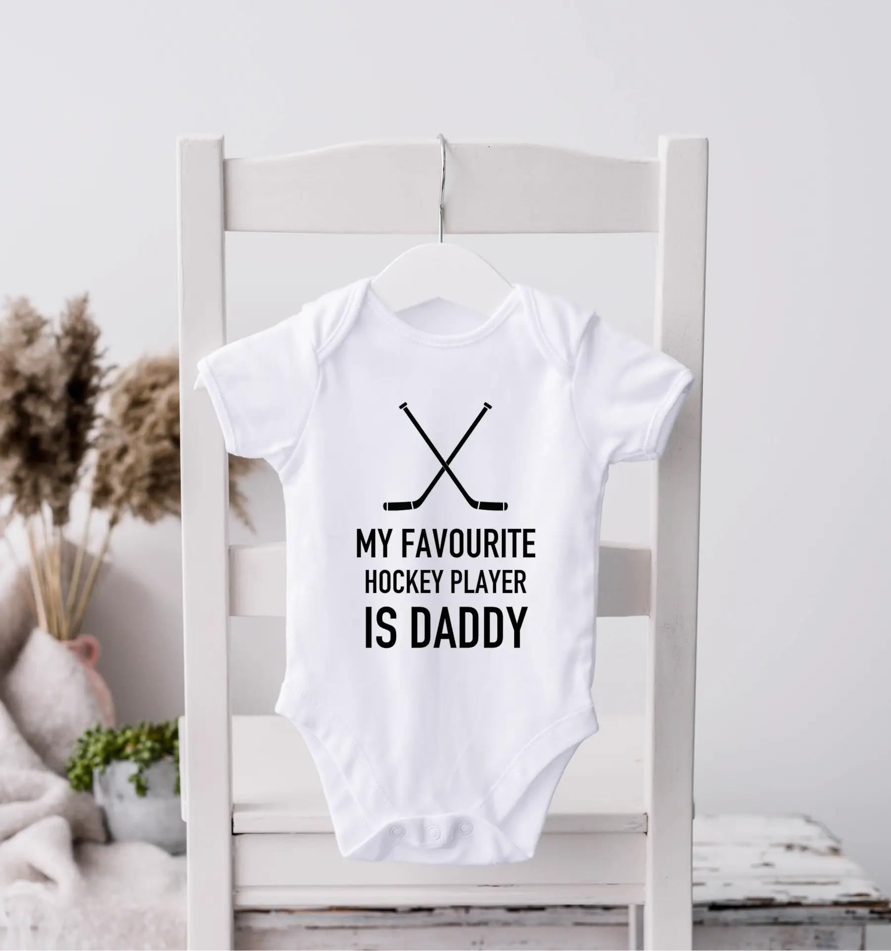White baby onesie with 'My favourite hockey player is Daddy' and crossed hockey sticks design on a hanger against a wooden chair with pampas grass – Maple Stick Hockey product.