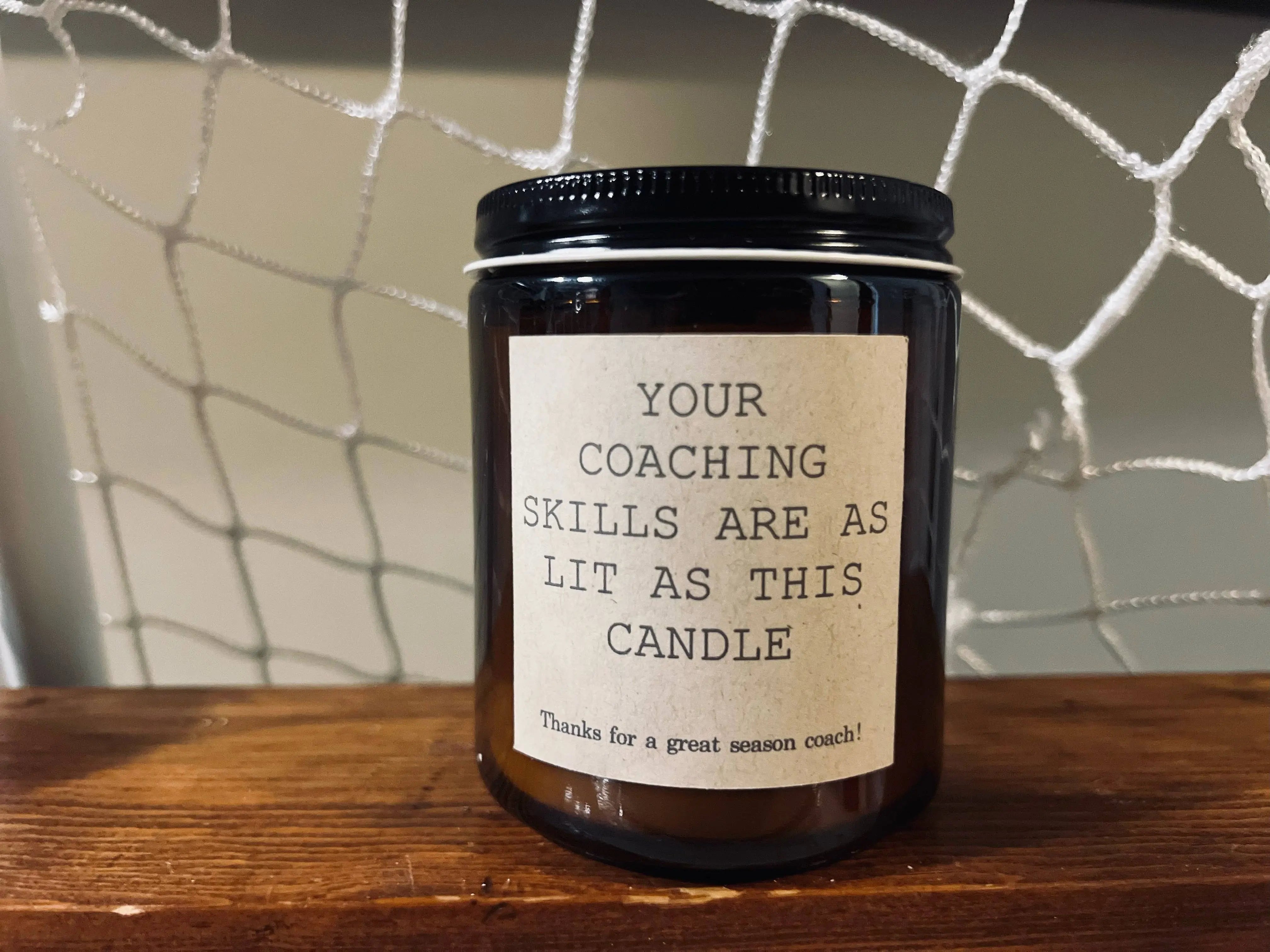 Brown glass candle with 'Your coaching skills are as lit as this candle' label on a wooden surface against a hockey net – Maple Stick Hockey product.