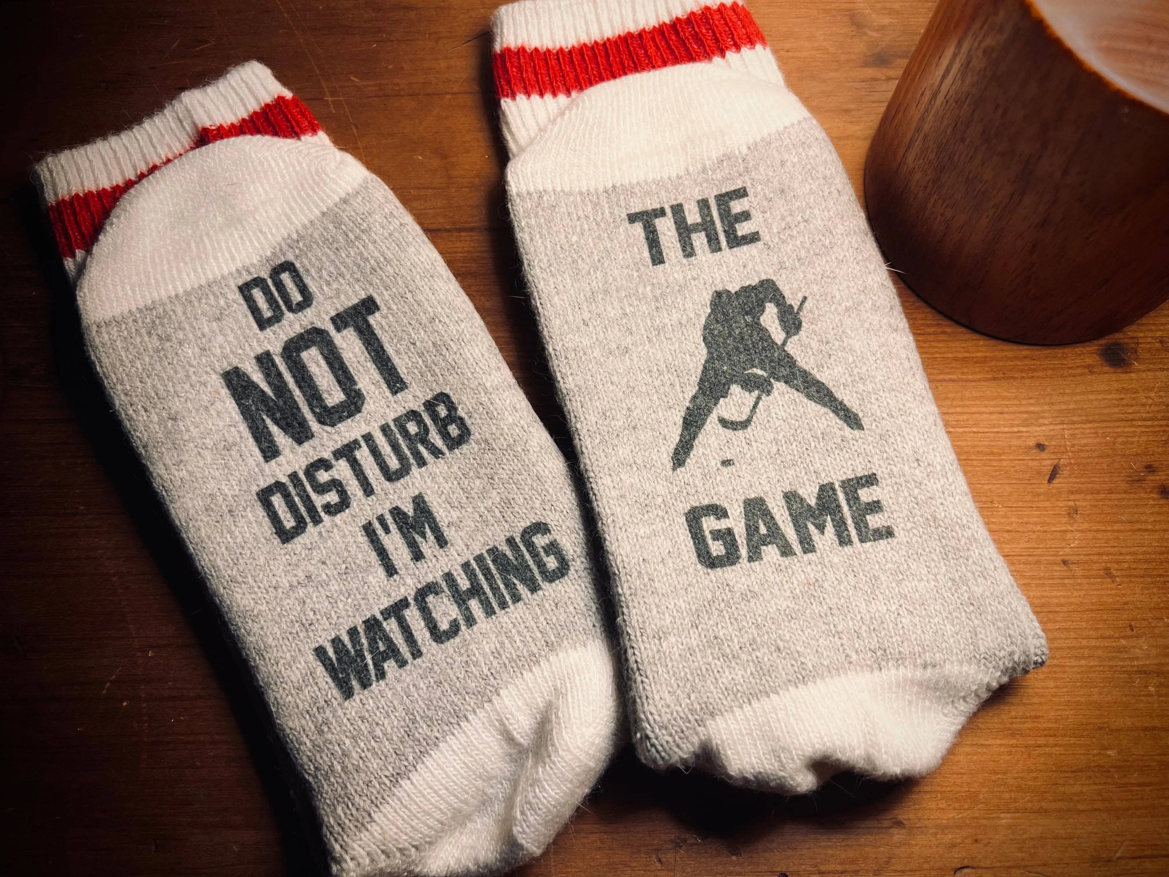 Pair of grey hockey socks: one sock says 'Do Not Disturb I'm Watching Hockey' and the other shows a hockey player silhouette with 'The Game' printed – Maple Stick Hockey product.