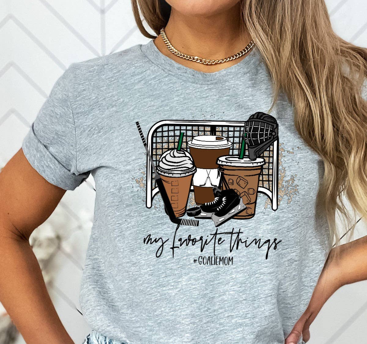Woman wearing grey 'Goalie Mom' T-shirt with 'My favourite things' graphic featuring coffee cups, skates and hockey gear – Maple Stick Hockey product.