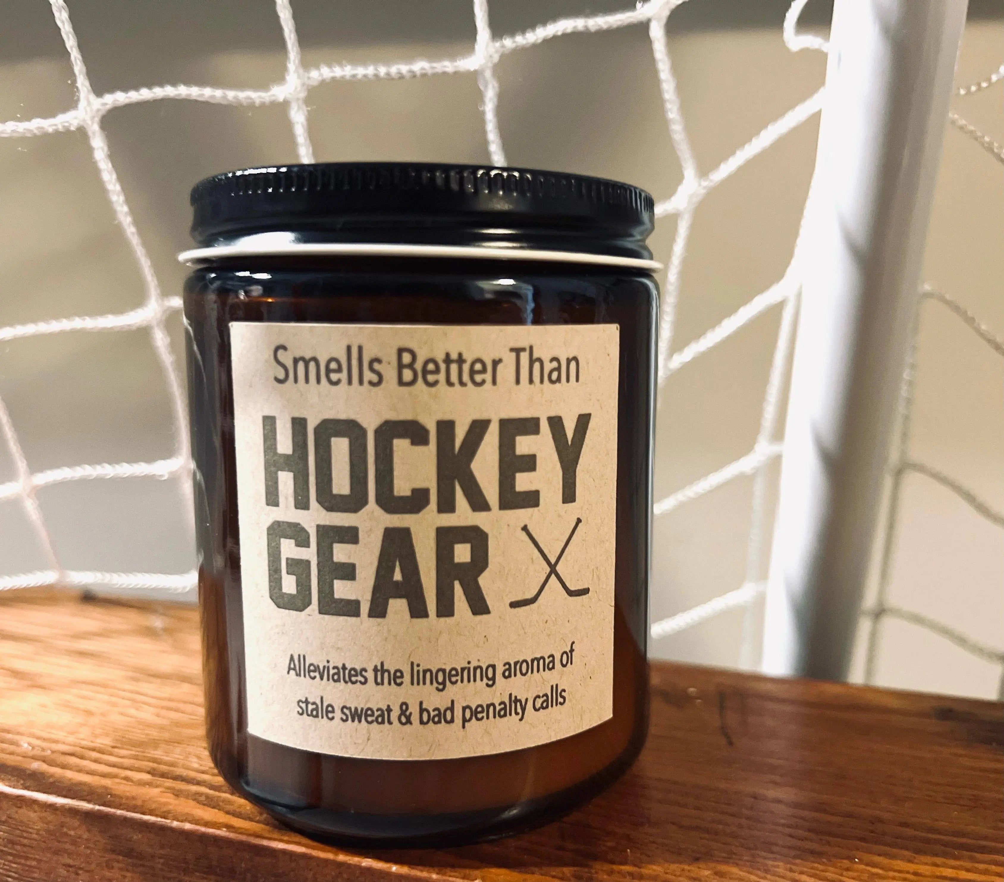 Brown glass candle labeled 'Smells better than hockey gear' with gold lid on a wooden surface in front of a hockey net – Maple Stick Hockey product.